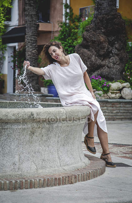 Feliz alegre mujer juguetona sentada en la fuente y dibujando agua en las manos en la calle Marbella - foto de stock