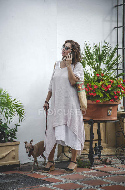 Оптимистичная модная счастливая женщина ходит по узкой улице в Марбелье, держа собаку на поводке, разговаривая по телефону — стоковое фото