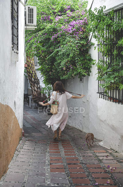 Back view of woman in casual summer dress with bag walking down in Marbella street with Italian Greyhound dog on leash — Stock Photo