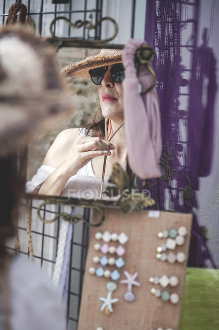 Стильна впевнена жінка в сонцезахисних окулярах дивиться в дзеркало для перевірки солом'яного капелюха з шнуром на міському ринку — стокове фото