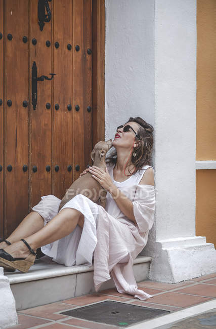 Vista lateral de la mujer con estilo sonriente en vestido de verano sosteniendo perro mientras está sentado en la puerta - foto de stock