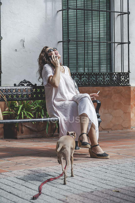 Fröhliche positive Frau mit Zigarette telefoniert und lacht, während sie sich bei einem Spaziergang mit Hund auf einer Bank ausruht — Stockfoto