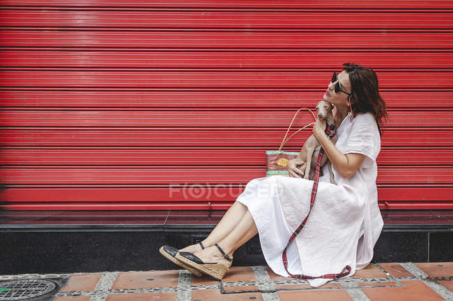 Vista lateral da mulher alegre satisfeita abraçando o cão bonito enquanto relaxa perto da parede listrada vermelha urbana na província de Espanha — Fotografia de Stock