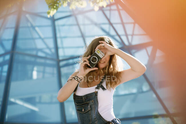 Jeune femme enthousiaste capturant moment prendre des photos sur la caméra sur fond d'architecture en verre — Photo de stock