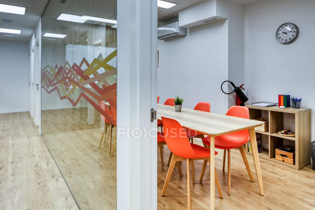 Светлый коридор с деревянным полом среди стеклянных стен светлых современных уютных офисных конференц-зон с удобными оранжевыми стульями за большими деревянными столами — стоковое фото