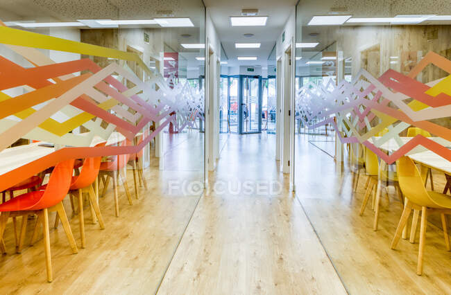 Corredor de luz con suelo de madera entre paredes de vidrio de luz modernas y acogedoras zonas de conferencias de oficina con cómodas sillas naranjas y amarillas en grandes mesas de madera - foto de stock