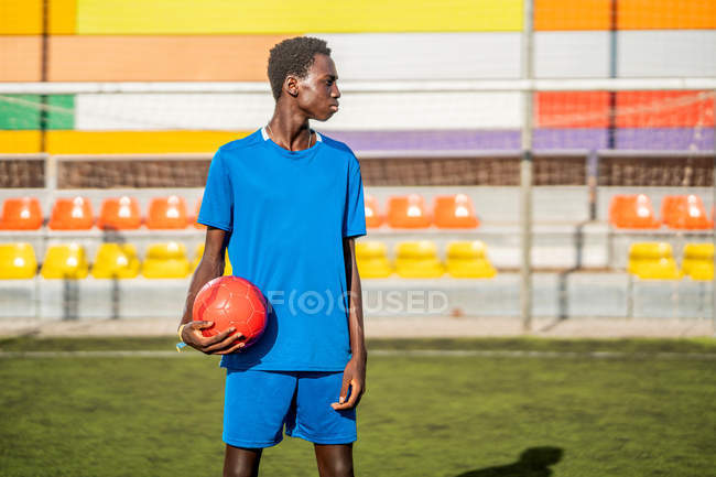 Jugador de fútbol negro con pelota de pie en el estadio y mirando hacia otro lado - foto de stock