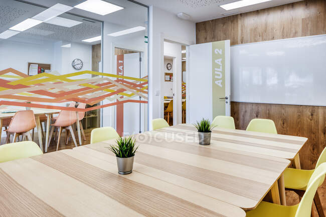 Современный интерьер светлого просторного офиса, окруженного стеклянной стеной с удобными желтыми стульями и серыми барными стульями за деревянными столами — стоковое фото