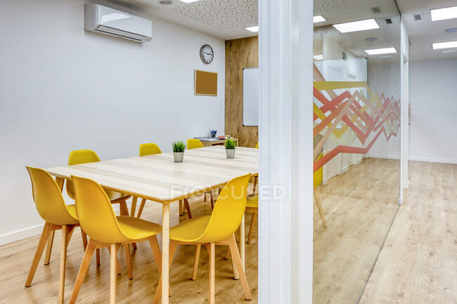 Светлый коридор с деревянным полом среди стеклянных стен светлых современных уютных офисных конференц-зон с удобными желтыми стульями за большими деревянными столами — стоковое фото