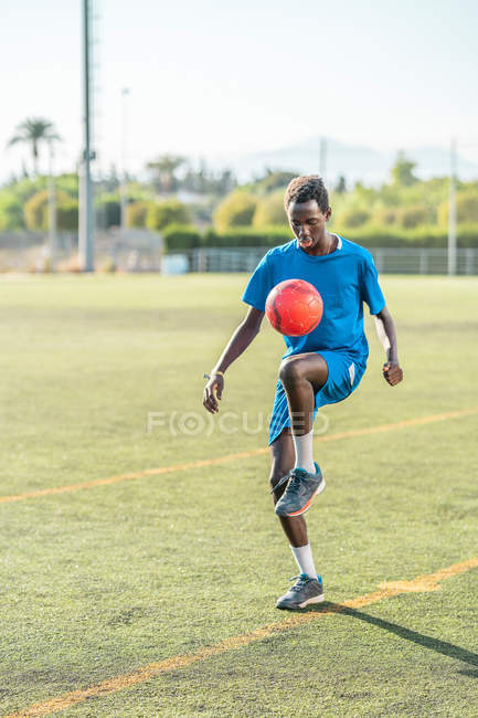 Этнический подросток в синей спортивной одежде жонглирует красным мячом во время тренировки на футбольном поле — стоковое фото