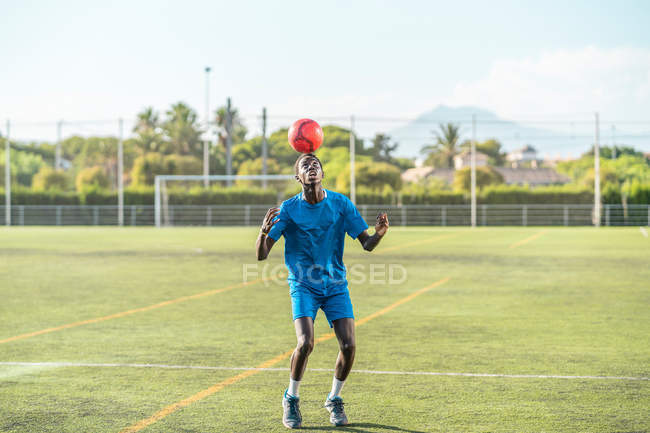 Magro adolescente preto malabarismo bola de futebol na cabeça no campo verde — Fotografia de Stock