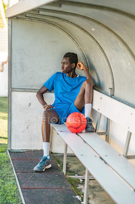 Longitud completa de adolescente afroamericano con pelota de fútbol sentado en el banco y mirando hacia otro lado durante el entrenamiento - foto de stock