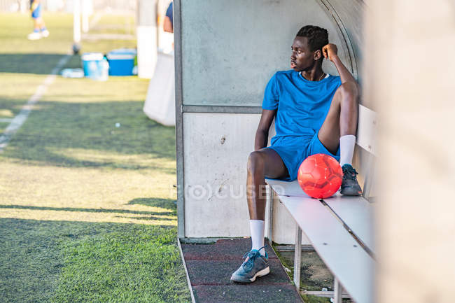 Longitud completa de adolescente afroamericano con pelota de fútbol sentado en el banco y mirando hacia otro lado durante el entrenamiento - foto de stock