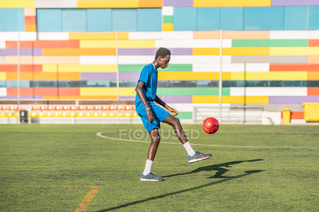Этнический подросток жонглирует футбольным мячом на зеленом поле — стоковое фото