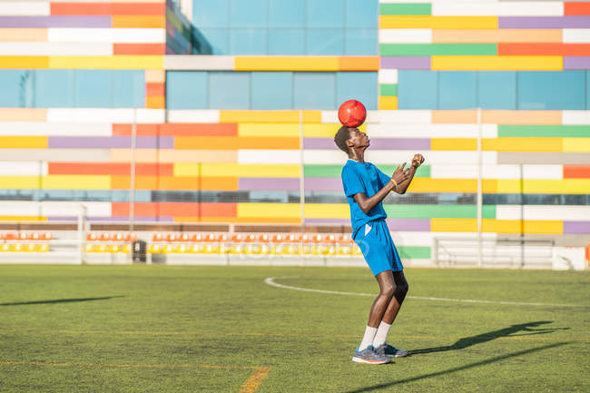 Africano americano adolescente em azul uniforme saltando bola brilhante na cabeça durante o treinamento no campo de futebol — Fotografia de Stock