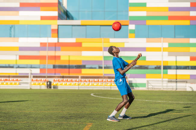 Adolescent afro-américain en uniforme bleu rebondissant balle brillante sur la tête pendant l'entraînement sur le terrain de football — Photo de stock