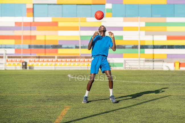 Афроамериканский подросток в синей форме, прыгающий яркий мяч на голове во время тренировки на футбольном поле — стоковое фото