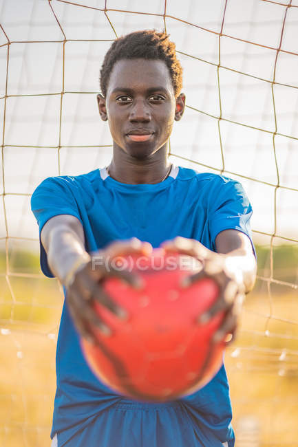 Adolescente negro en camiseta azul llevando bola roja en brazos extendidos y mirando a la cámara contra la red de gol - foto de stock