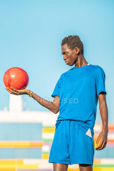 Черный подросток держит красный футбольный мяч против безоблачного неба — стоковое фото