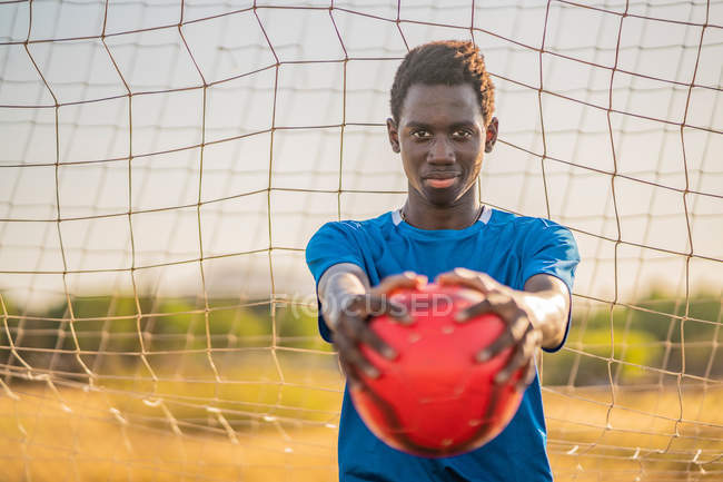 Adolescente preto em camiseta azul carregando bola vermelha em braços estendidos e olhando para a câmera contra a rede de gol — Fotografia de Stock
