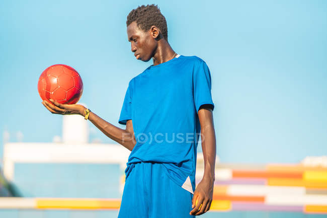 Nero adolescente tenendo rosso pallone da calcio contro nuvoloso cielo — Foto stock