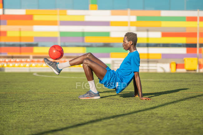 Вид сбоку на этнического футболиста, откинувшегося назад и жонглирующего мячом на ногах во время тренировки на стадионе — стоковое фото