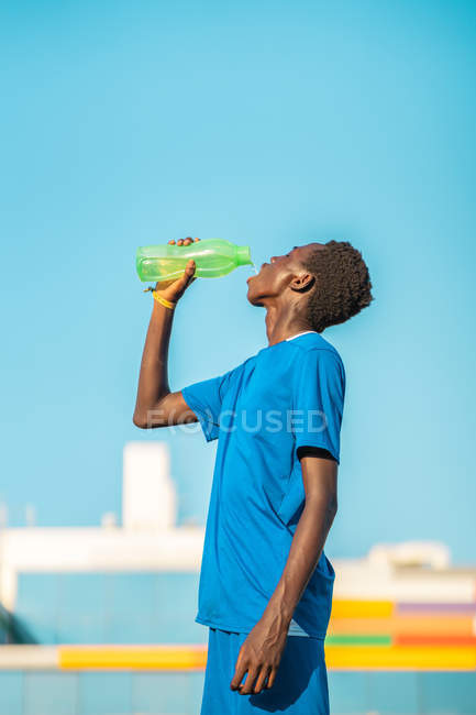 African American teenager with ball drinking fresh water during football training on sunny daytime — Stock Photo