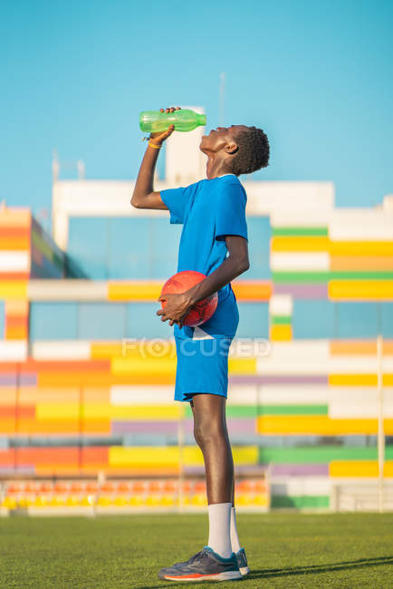 Афроамериканский подросток с мячом, пьющий пресную воду во время футбольных тренировок в солнечный день — стоковое фото