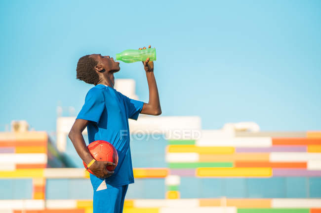 Adolescente afro-americano com bola bebendo água doce durante o treinamento de futebol no dia ensolarado — Fotografia de Stock