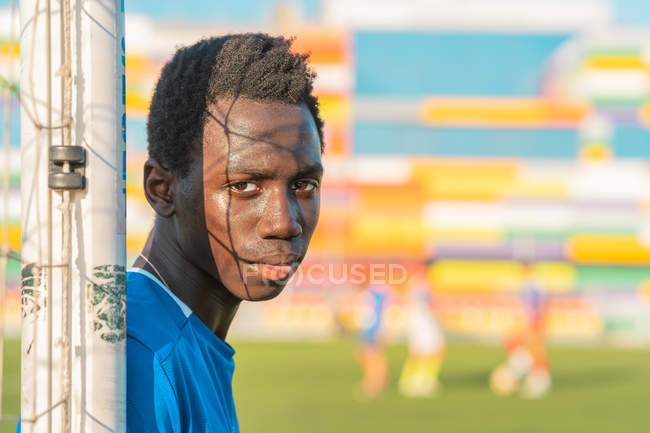 Adolescente étnico olhando na câmera enquanto se inclina na rede no fundo turvo do campo de futebol — Fotografia de Stock