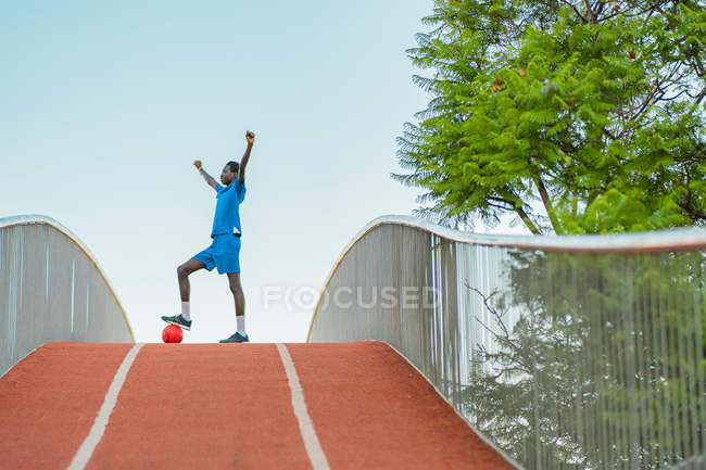 Полная длина черного подростка в синей спортивной одежде, наступающего на футбольный мяч и поднимающего руки во время тренировки на улице — стоковое фото
