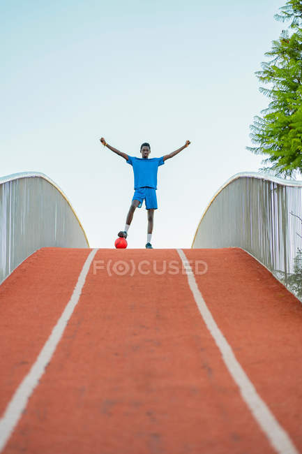 Full length of black teenager in blue sportswear stepping on football ball and raising arms during training on street — Stock Photo