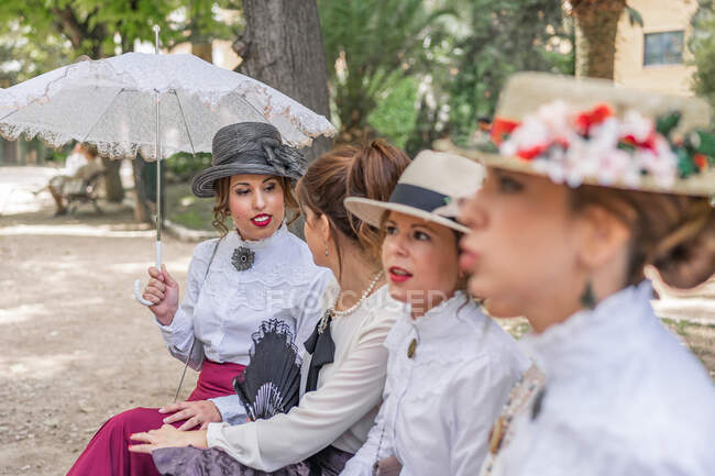 Группа женщин в винтажных платьях разговаривает друг с другом, сидя на скамейке в парке — стоковое фото