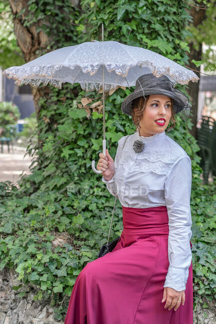 Елегантна жінка в ретро-сукні і з мереживною парасолькою посміхається і дивиться геть, сидячи біля дерева в парку — стокове фото
