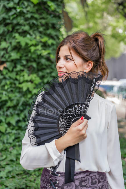 Jovem mulher alegre em roupa vintage elegante segurando ventilador preto e olhando para longe enquanto está em pé no jardim verde — Fotografia de Stock