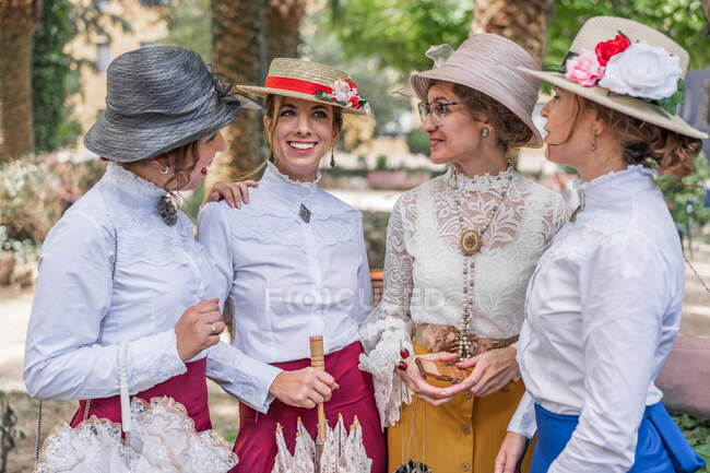 Группа веселых женщин в старомодных платьях, улыбающихся и разговаривающих друг с другом, стоя в парке вместе — стоковое фото