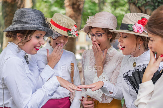 Mulher alegre mostrando anel de noivado para surpreendeu amigos do sexo feminino em vestidos vintage e chapéus enquanto passava o tempo no parque — Fotografia de Stock