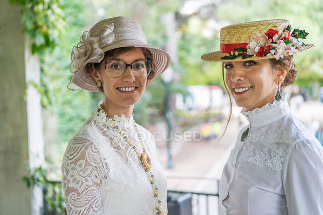 Frauen in Retro-Hüten und Blusen lächeln und blicken in die Kamera, während sie vor verschwommenem Hintergrund des Parks stehen — Stockfoto