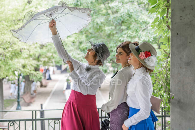 Весела жінка в ретро-сукні посміхається і відкриває мереживні парасольки, стоячи на терасі в саду біля друзів-жінок — стокове фото