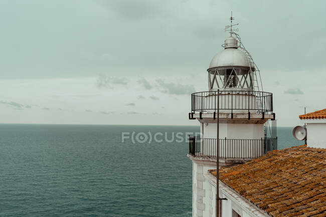 D'en haut le phare blanc est resté à côté du bâtiment avec le toit carrelé rouge sur la rive de la mer avec l'eau sombre calme et le ciel nuageux gris — Photo de stock