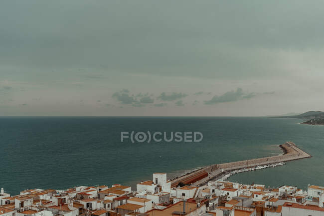 Aerial view of sea port with tightly stayed buildings with red roofs and pier with boats on coast with dark water and grey cloudy sky — Stock Photo