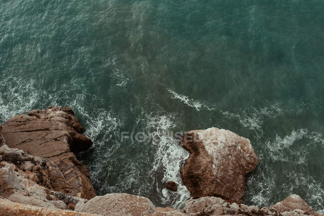 Aerial view of sea waves hitting rocky cliff with splash and foam on seashore with deep dark water — Stock Photo