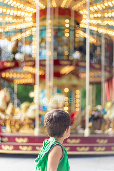 Vista posteriore del bambino piccolo in abiti casual verdi guardando lontano e contemplando mentre in piedi da solo contro colorato girevole allegro andare in giro nel parco divertimenti durante il giorno — Foto stock