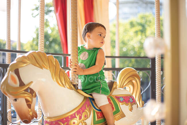 Bambino curioso pensieroso in abiti casual verdi guardando lontano mentre cavalca favoloso cavallo di carnevale da solo nel parco divertimenti nella giornata di sole — Foto stock
