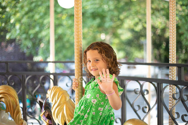 Sonriente niño pensativo en ropa casual verde mirando hacia otro lado y saludando la mano mientras monta fabuloso pony de carnaval en el parque de atracciones - foto de stock