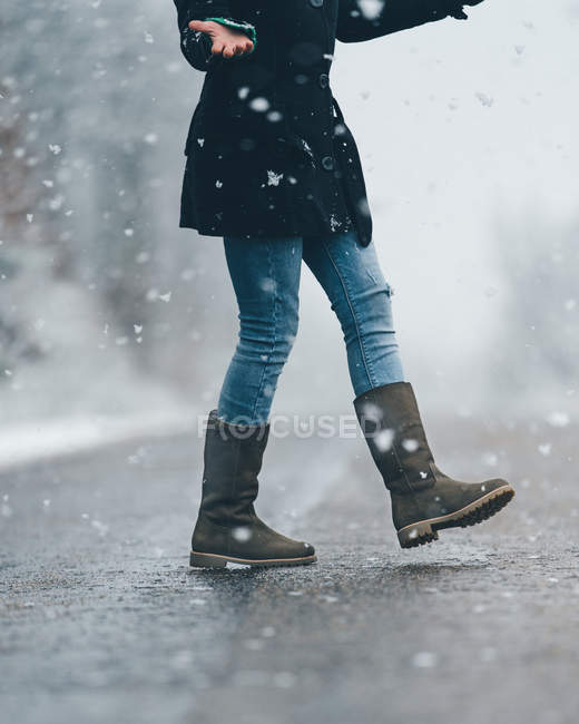 Visão lateral de seção baixa da menina cruzando estrada rural vazia na neve e clima de inverno sombrio — Fotografia de Stock