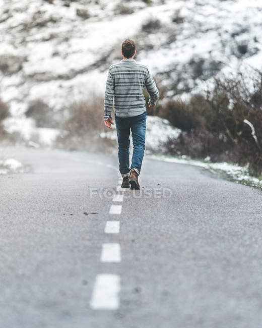 Vista trasera del hombre caminando por la carretera del campo con colinas cubiertas de nieve en un clima sombrío y nublado - foto de stock