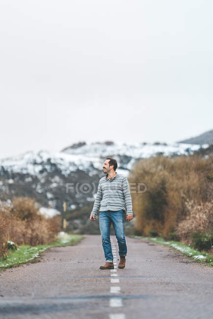 Взрослый мужчина идет по проселочной дороге с холмами, покрытыми снегом в мрачную облачную погоду — стоковое фото