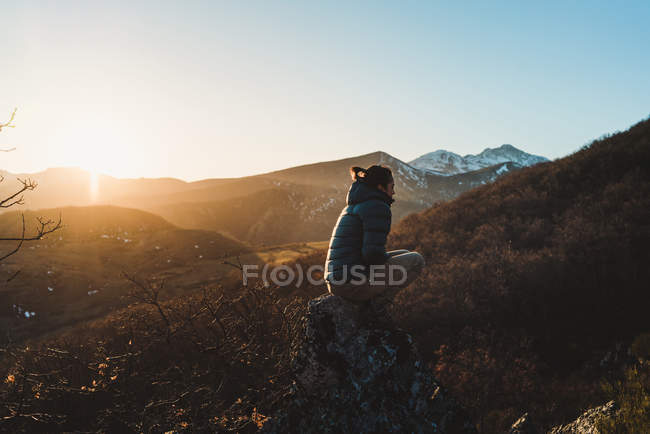 Вид сбоку молодой женщины в активной одежде, сидящей на склоне горы на красивом осеннем закате — стоковое фото