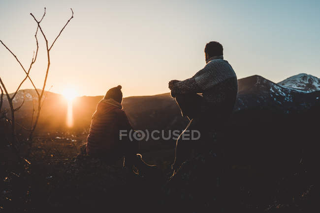 Silhueta de homem e mulher relaxados sentados em backlit no topo da montanha ao pôr do sol — Fotografia de Stock